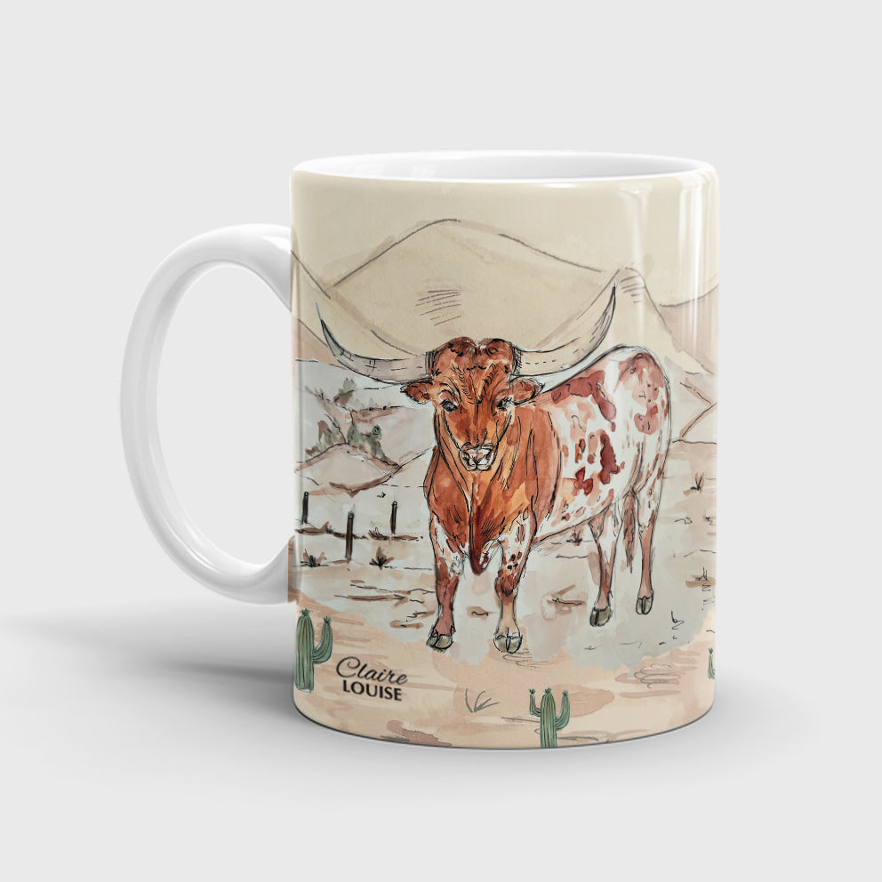 A white ceramic mug featuring a painted illustration of a longhorn steer in a desert landscape.