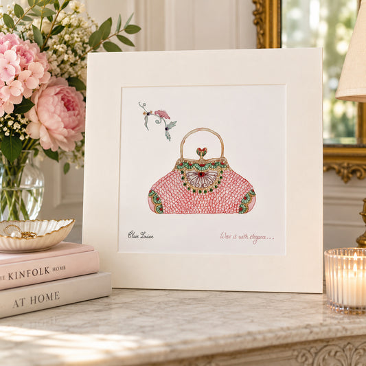 A framed art print featuring a pink embroidered handbag illustration with green and gold details, styled on a marble surface next to a vase of pink flowers and books.