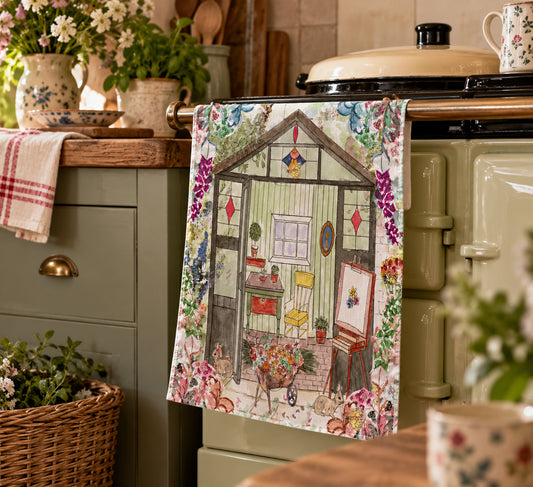 A kitchen towel featuring a vintage-style illustration of a cozy greenhouse with flowers and animals, hanging on a sage green stove in a rustic kitchen setting.
