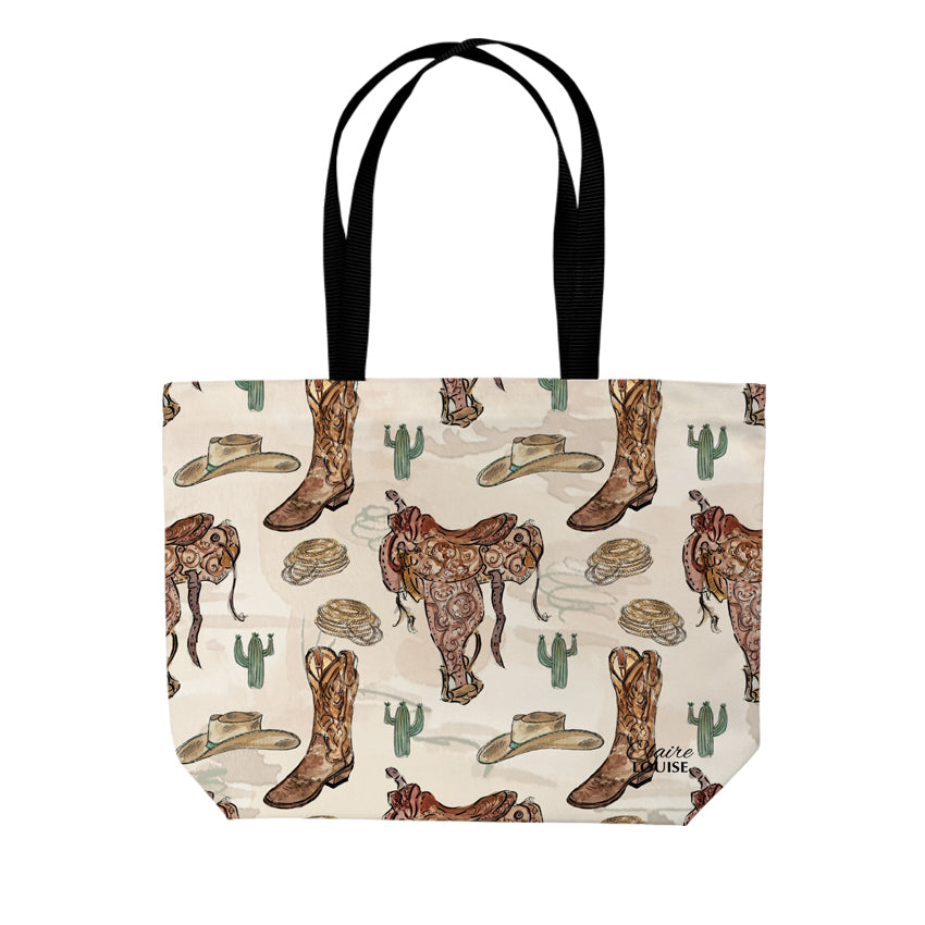A tote bag with a western-themed print featuring cowboy boots, hats, cacti, and a saddle on a cream background, with black shoulder straps, shown against a plain white background.