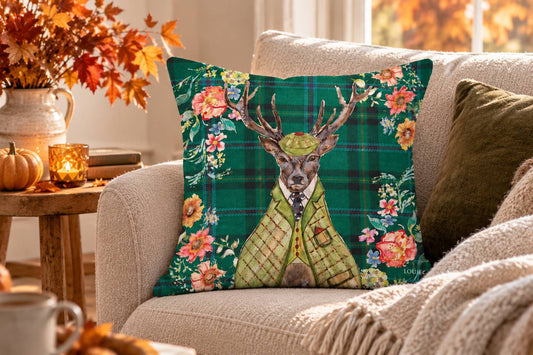 A decorative throw pillow featuring a deer in a green plaid vest and hat surrounded by autumn flowers, styled on a beige sofa in a cozy living room setting.
