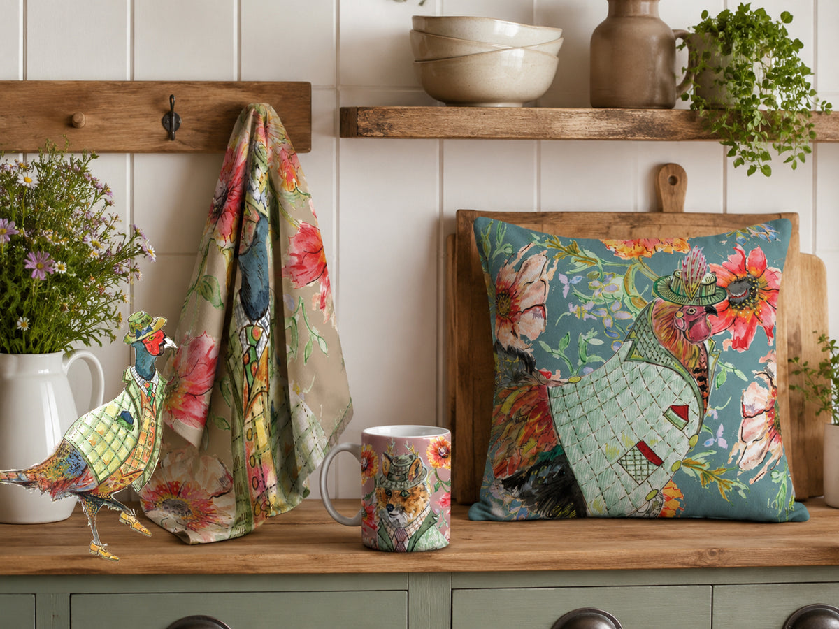 A decorative throw pillow with a colorful bird and floral print sits on a wooden kitchen shelf next to a matching printed mug and a draped fabric piece. The scene is styled with ceramic pitchers, potted plants, and a wooden cutting board against a white shiplap wall.