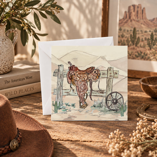 A square art print or card featuring a western illustration of a brown leather saddle in front of a wooden fence, displayed on a rustic wooden table with a white envelope behind it. The scene is styled with a cowboy hat, dried flowers, books, a ceramic pitcher, and framed western art in warm natural light.