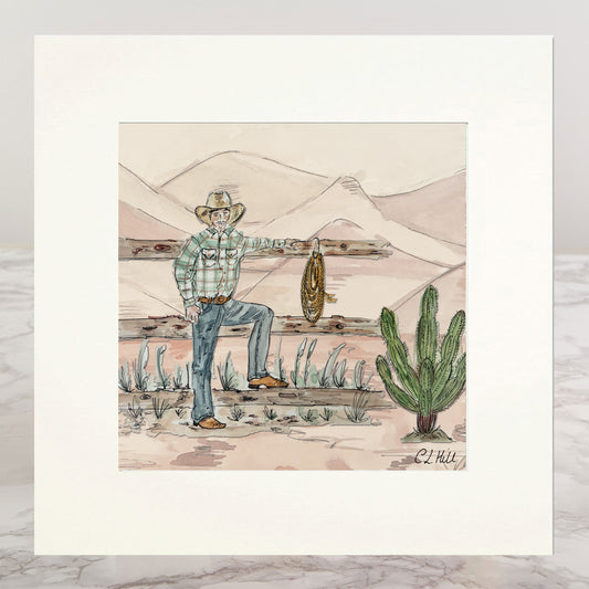 A matted art print featuring a hand-drawn western illustration of a cowboy in a plaid shirt and jeans, holding a lasso, with mountains, a fence, and a cactus in the background.