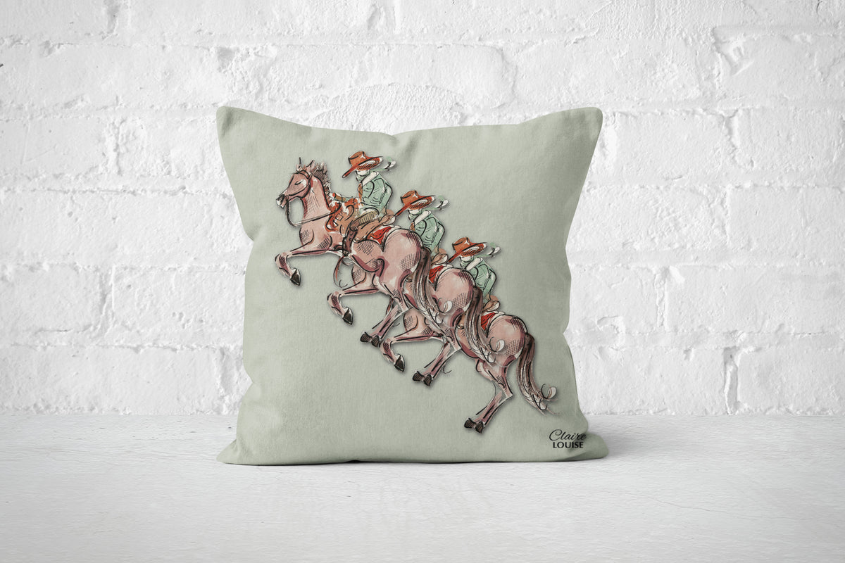A square sage green throw pillow featuring a printed illustration of three cowboys.