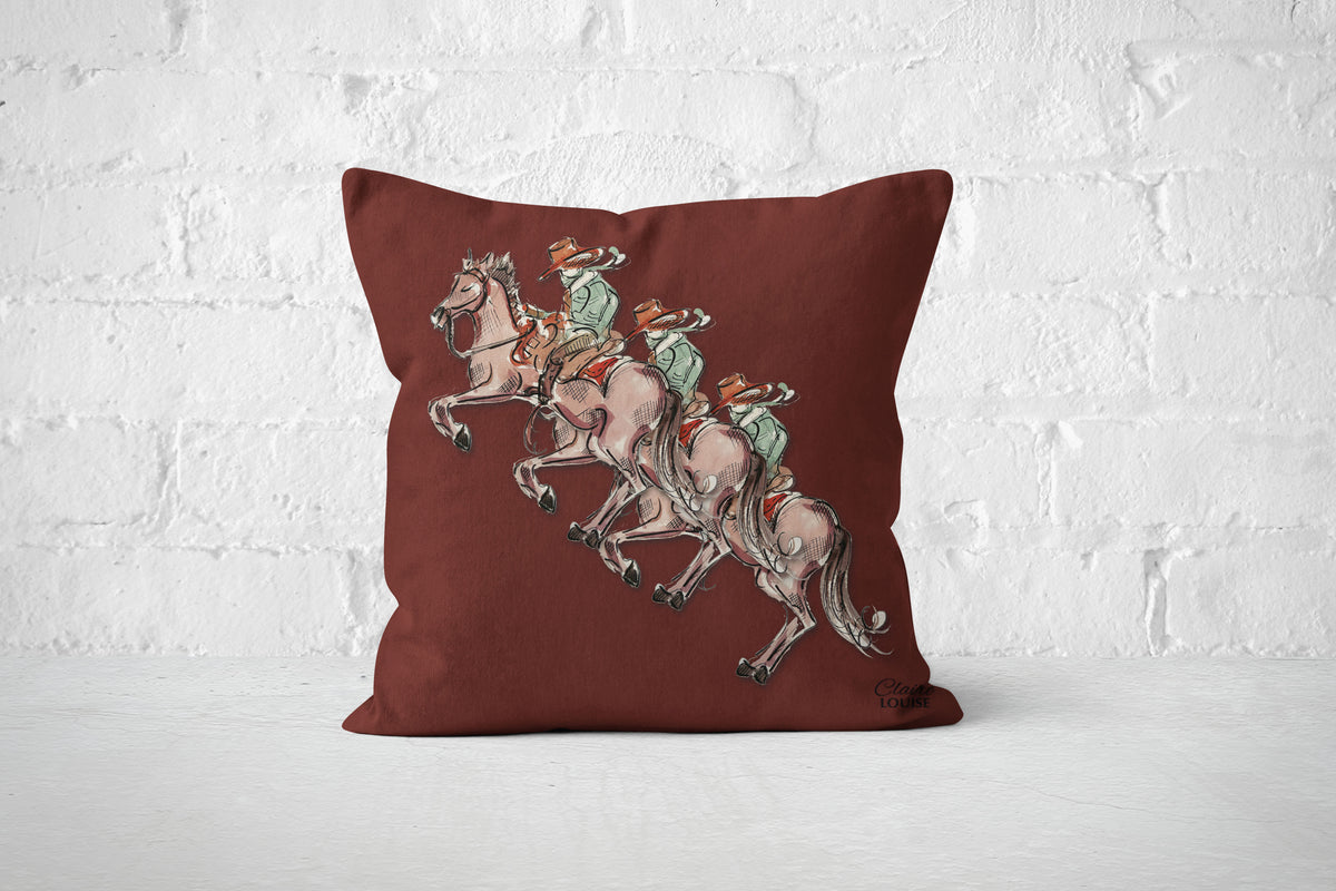 A square throw pillow in a deep burgundy red color, featuring a printed graphic of three cowboys.
