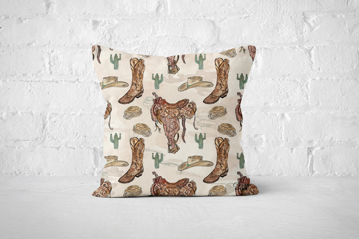 A square throw pillow with a western-themed print featuring brown cowboy boots, a brown steer, green cacti, and beige hats on a cream background, set against a white brick wall backdrop.