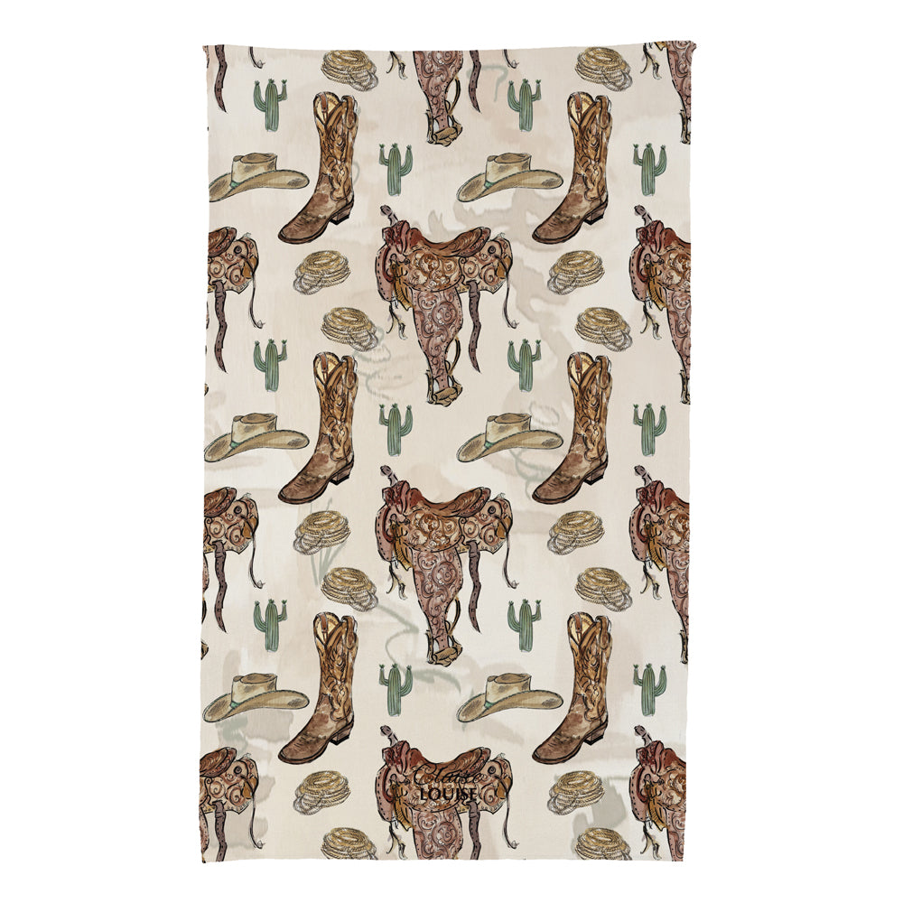 A rectangular fabric item, likely a towel or blanket, featuring a western-themed print with brown cowboy boots, saddles, hats, cacti, and skulls on a light beige background.