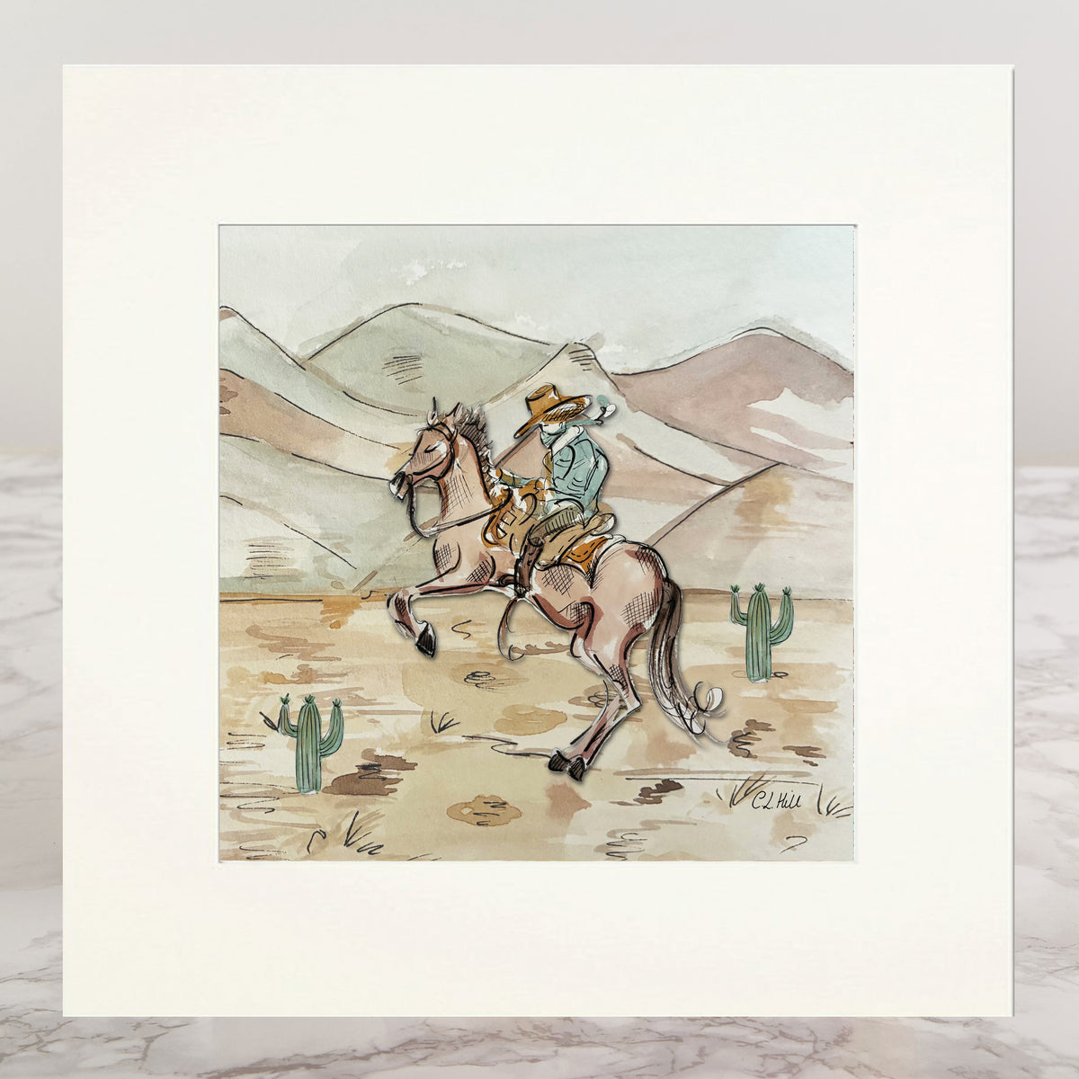 A matted art print featuring a western illustration of a cowboy riding a horse across a desert landscape with mountains and cacti, displayed on a light marble surface.