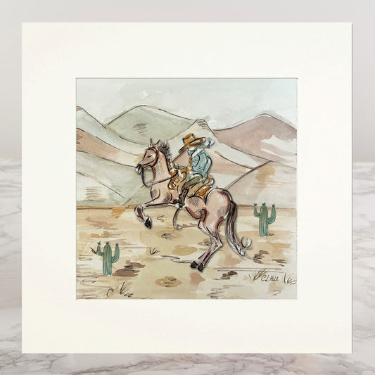 A matted art print featuring a western illustration of a cowboy riding a horse across a desert landscape with mountains and cacti, displayed on a light marble surface.